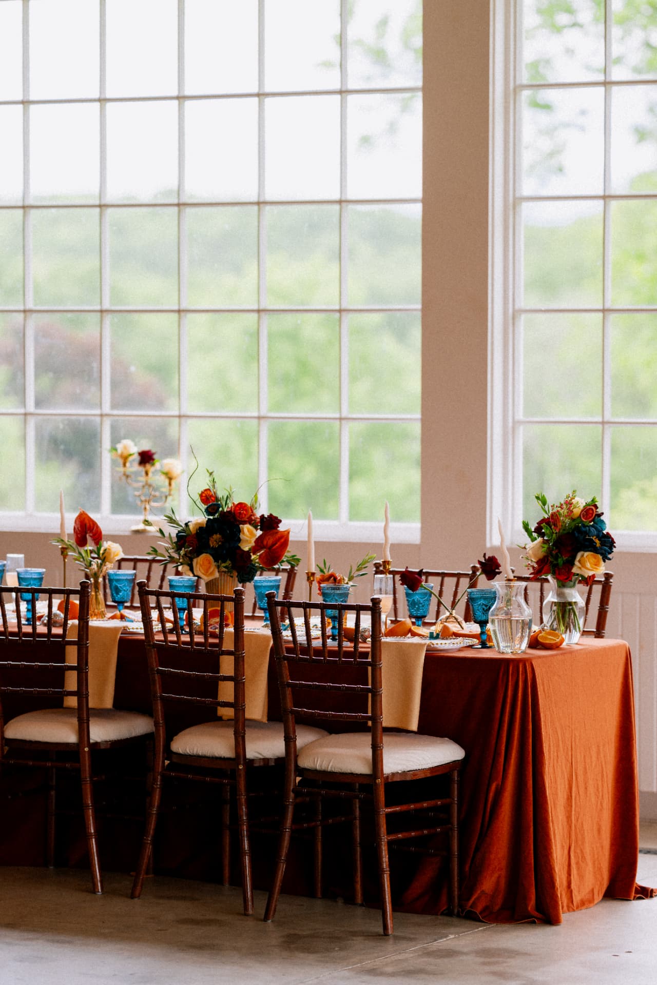 A long table with rust linens, blue glassware, and colorful florals in front of tall windows