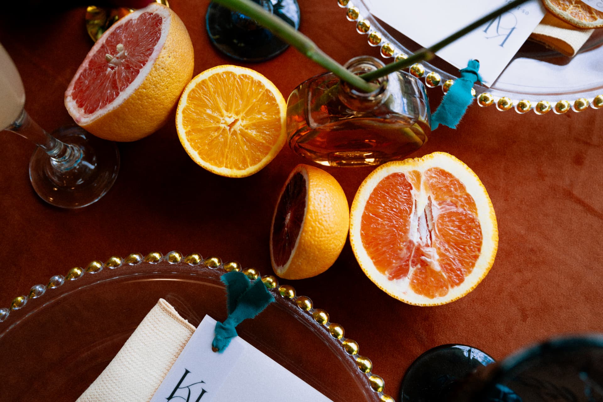 Halved citrus, amber glass, and teal ribbon on a rust velvet tablecloth