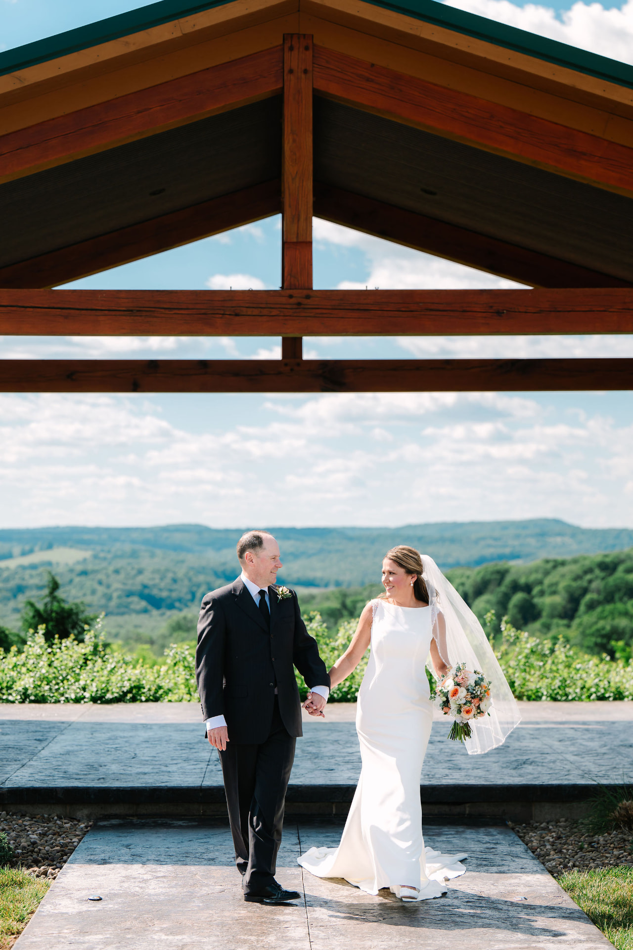 Greg and Sonnie holding hands beneath a timber pavilion with hills behind them